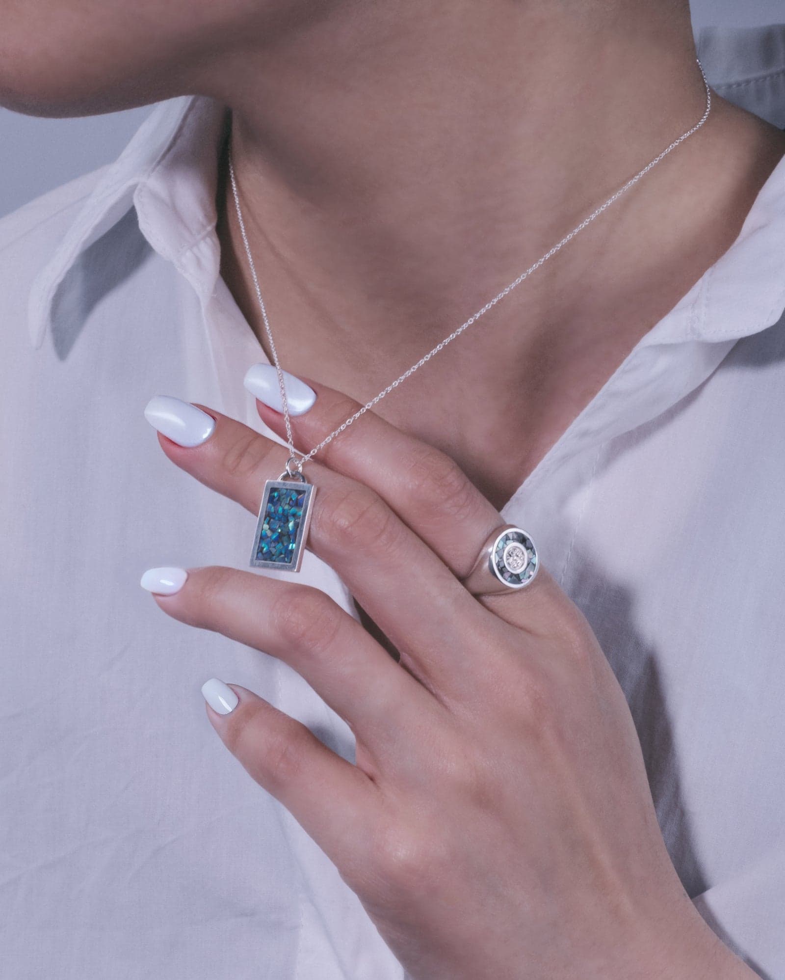 A hand wearing an ERNA signet ring and holding a rectangle pendant against a white shirt